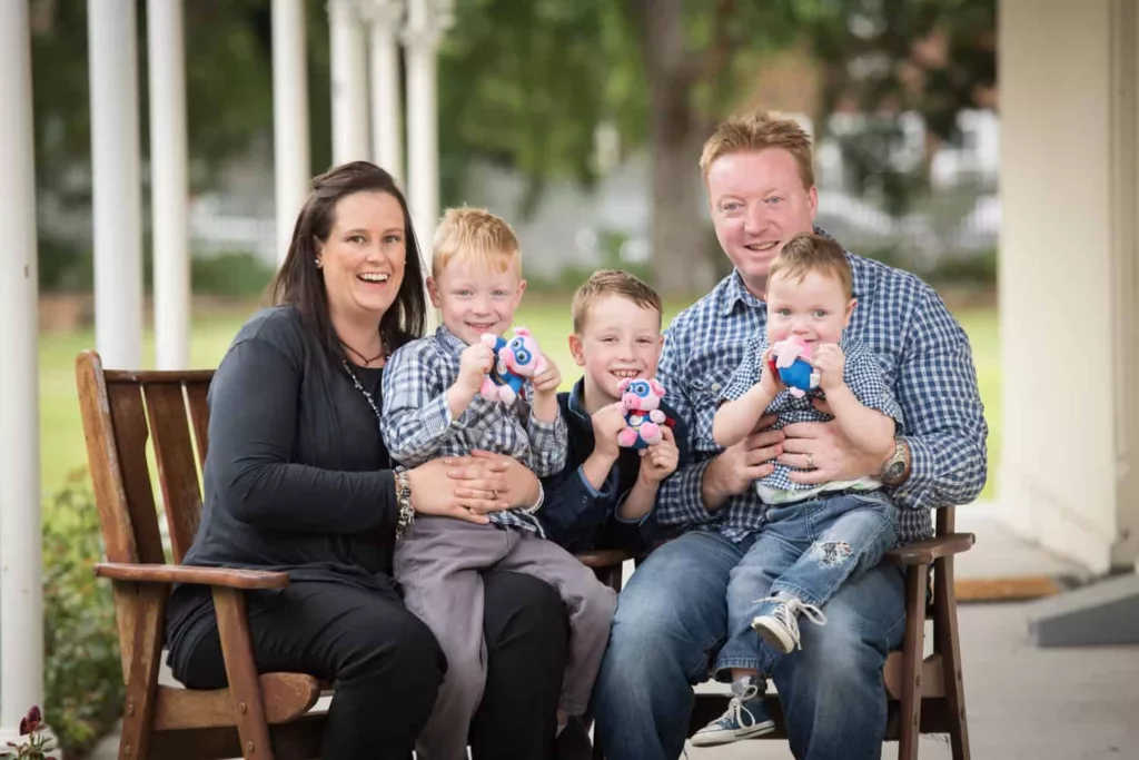 Two adults and three children are sitting outdoors. The children are holding pig shaped toys and everyone is smiling at the camera.