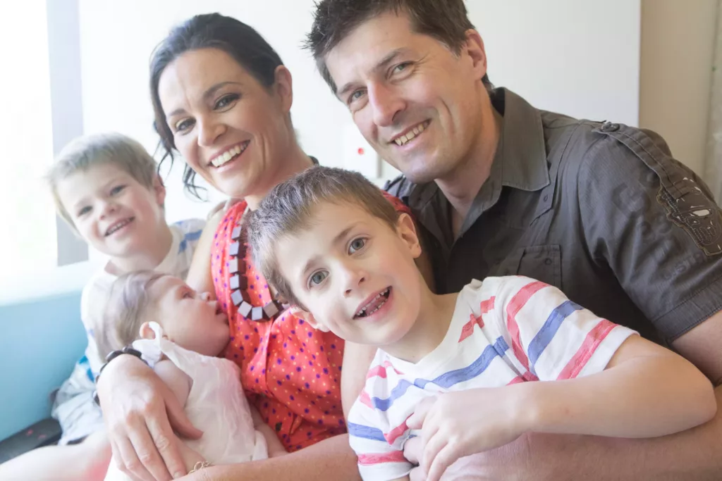 A family of two adults, two young children and a baby are seated inside, they are smiling at the camera.