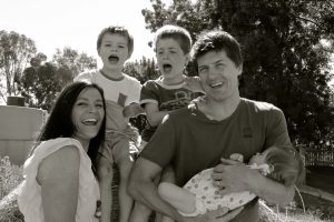 A family of two adults, two children and a baby are outdoors with trees in the background, they are laughing and smiling for the camera.