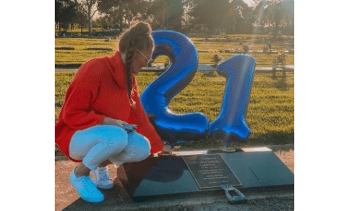 A lady is kneeling by a gravestone, there are balloons tied to it of a 2 and a 1 for his 21st birthday. The sun is shining.