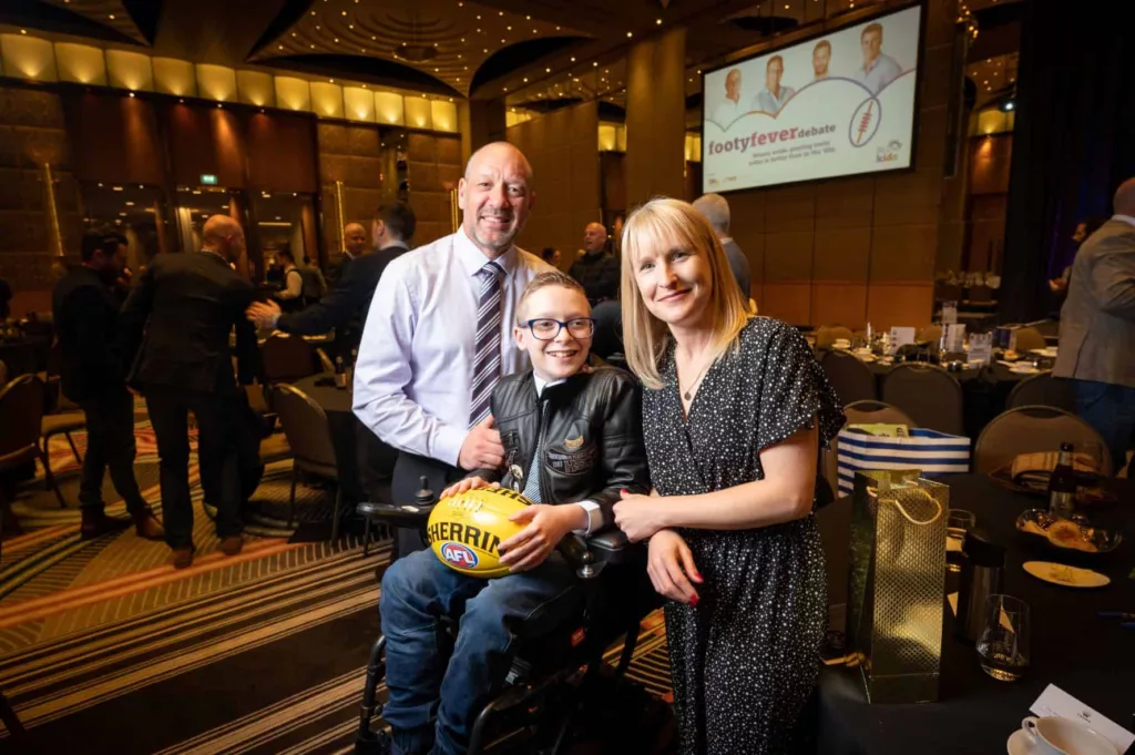 A boy is sitting in his wheelchair, holding a football. His parents are on either side, smiling at the camera.