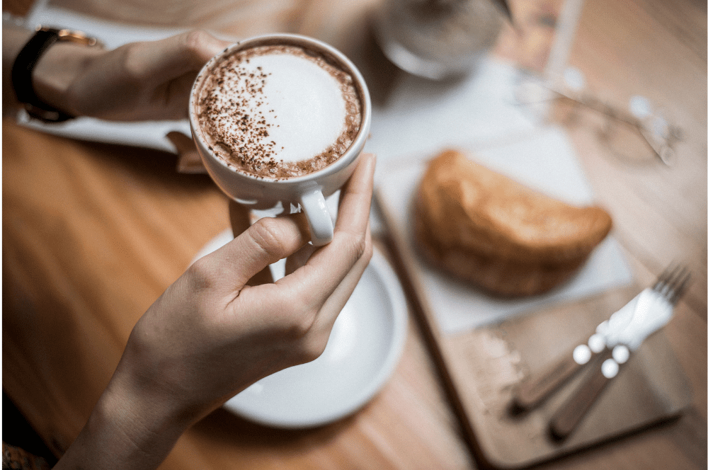 Image of someone's hands holding a cup of coffee. There is a pastry on the table in the background.