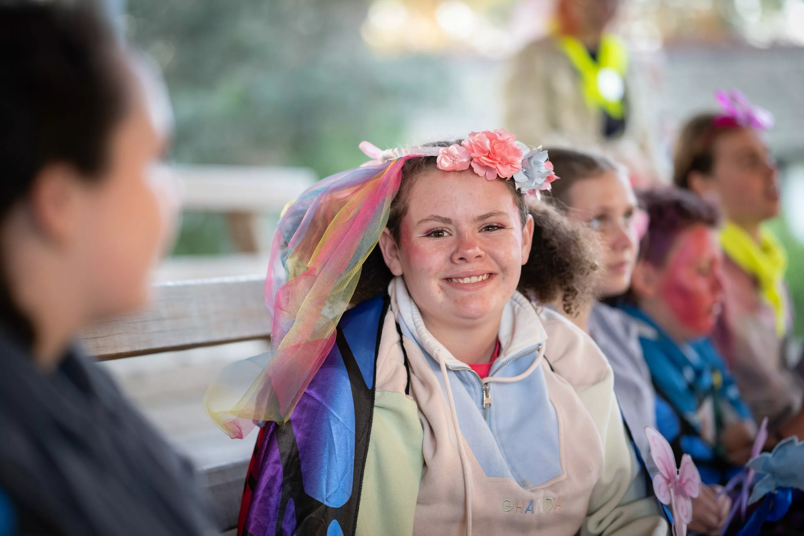 A young adolescent woman is wearing a costume veil, and bright coloured clothing. She is sitting outdoors smiling at the camera.