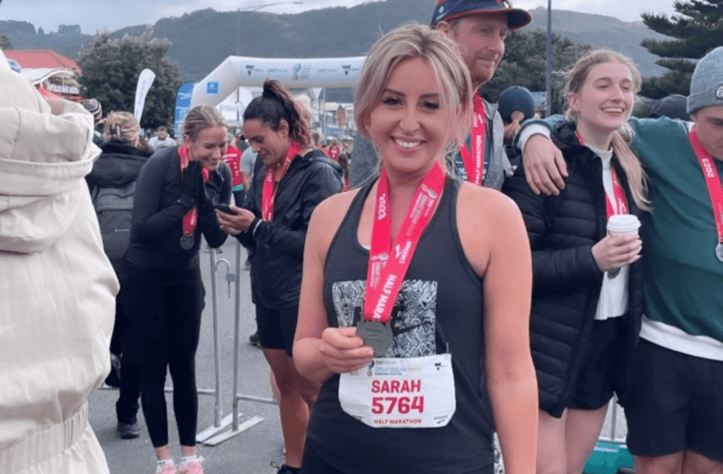 A young woman is standing outdoors, she is wearing running clothes and is showing a medal around her neck while smiling at the camera. There are other runners and spectators in the background.