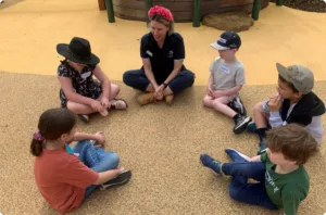A group of children and an adult are sitting in a circle outdoors playing games