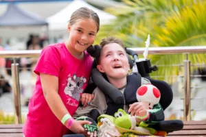 A girl is smiling at the camera as she stands next to a boy sitting in a wheelchair
